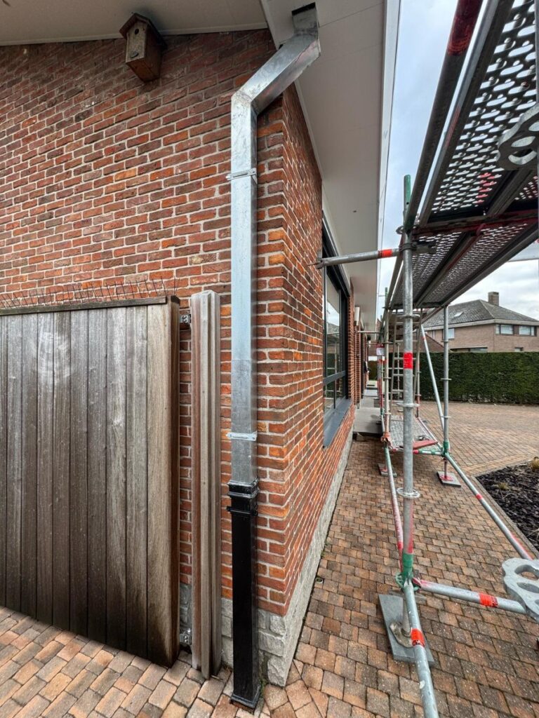 Nieuwe zinken regenpijp langs een bakstenen gevel tijdens dakwerken